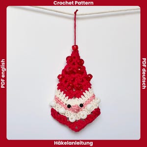 Peut inclure: Ornement de sapin de Noël fait à la main au crochet. L'arbre rouge est orné de perles rouges et d'accents blancs et roses. L'ornement a une ficelle pour l'accrocher et est sur fond blanc. Le texte "Crochet Pattern" et "Häkelanleitung" sont visibles.