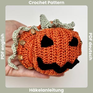 Könnte beinhalten: Ein gehäkelter orangefarbener Kürbis mit einem geschnitzten Gesicht, schwarzen Augen und Mund sowie einem grünen Stiel. Der Kürbis ist mit kleinen, braunen, samenartigen Verzierungen verziert. Der Text "Crochet Pattern" und "Häkelanleitung" sind sichtbar.