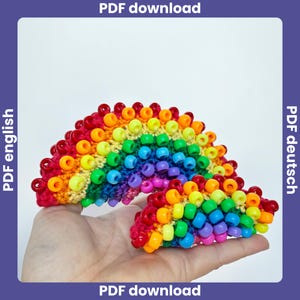 May include: A handmade rainbow-shaped craft item. The rainbow is made of colorful beads and yarn, with a vibrant array of colors including red, orange, yellow, green, blue, and purple. The craft item is held in a hand against a white background.