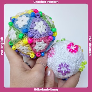 Puede incluir: Dos objetos esféricos de ganchillo con centros florales blancos rodeados de triángulos de hilo de colores. La esfera más grande muestra una paleta multicolor de amarillo, verde, azul y morado, mientras que la más pequeña es blanca. Ambas están adornadas con cuentas de plástico.