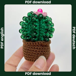 Peut inclure: Un cactus crocheté à la main avec des épines en perles vertes et une fleur rose, dans un pot en crochet marron. Le cactus est tenu dans une main sur fond blanc. L'image contient également le texte "PDF download".