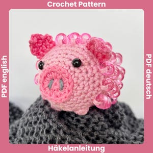 Pode incluir: Um porco rosa de crochê com olhos pretos e um focinho cinza. O porco tem enfeites rosa encaracolados ao redor da cabeça. A imagem contém o texto "Crochet Pattern" e "PDF deutsch".