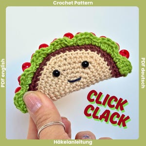 Könnte beinhalten: Ein gehäkeltes Taco-Spielzeug mit beiger Schale, brauner Füllung und einem grünen Salatblatt-Rand, der mit kleinen roten Perlen verziert ist. Das Taco hat zwei schwarze Perlenaugen und ein gesticktes Lächeln. Der Text CLICK CLACK ist in Rot und Grün.