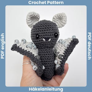 Pode incluir: Um morcego de crochê com fios cinza e preto, olhos grandes e pretos e pequenas presas brancas. As asas são detalhadas com contas cinza e brancas. A imagem inclui o texto "Crochet Pattern" e "PDF english".