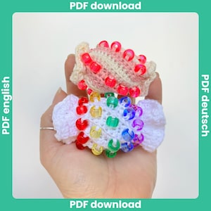 May include: Two handmade crochet hair scrunchies. One is white with a rainbow of colorful beads, and the other is cream and pink with red beads. The scrunchies are designed with a ruffled edge.