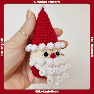 May include: A crocheted Santa Claus ornament with a red hat, white beard, and black eyes. The ornament is adorned with white beads and has the text "Crochet Pattern" and "Häkelanleitung".