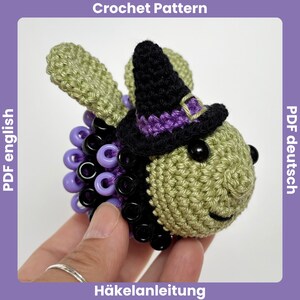 May include: A crocheted bee wearing a witch hat. The bee is olive green with black and purple accents. The witch hat is black with a purple band. The bee has black bead eyes and a smiling face. The image has the text "Crochet Pattern" and "PDF english".