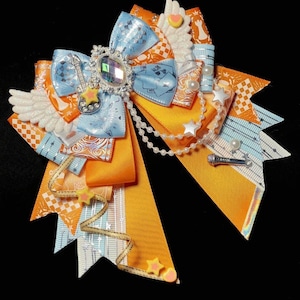 May include: A decorative hair bow featuring a large blue bow with a silver gem center and a guitar charm. The bow has orange and white patterned accents, white wings, and a pearl strand. Additional embellishments include star and heart shapes.