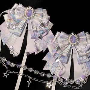 May include: Two decorative white and iridescent bows with purple accents and a silver chain with star charms. Each bow has a central oval-shaped jewel and is adorned with ribbons and star-shaped embellishments.