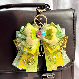 May include: A green and yellow bow keychain with a gold chain and a sparkly heart charm. The bow is made of multiple layers of ribbon and has a fluffy texture. The keychain is attached to a brown leather bag.