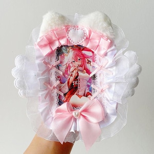 May include: A white and pink decorative accessory featuring a printed anime character. The design includes a pink bow, ribbons, and a heart-shaped detail. The top has fluffy white ears.