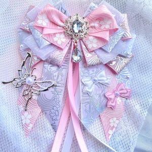 May include: A decorative bow in shades of pink, white, and lavender, featuring a central jeweled embellishment and a dangling crystal. The bow includes a silver butterfly accent and multiple ribbon streamers.