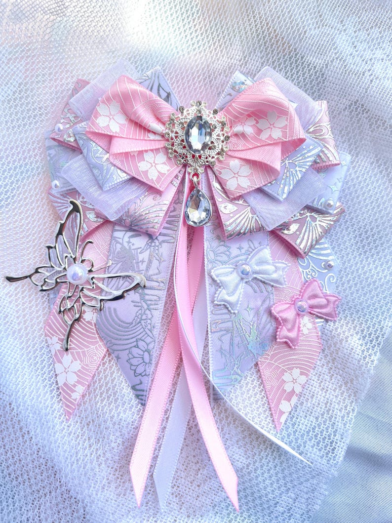 May include: A decorative bow in shades of pink, white, and lavender, featuring a central jeweled embellishment and a dangling crystal. The bow includes a silver butterfly accent and multiple ribbon streamers.
