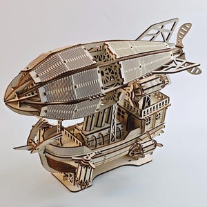 Puede incluir: Modelo de madera de un dirigible. El modelo presenta detalles intrincados, incluyendo el cuerpo, la góndola y las aletas traseras de la aeronave. Construido en madera clara, se exhibe sobre un fondo blanco, resaltando su diseño.