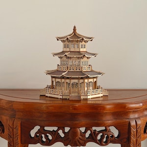 Puede incluir: Un modelo de madera de tres pisos de una pagoda que presenta detalles intrincados en las ventanas de celosía, múltiples aleros sobresalientes y una aguja central. La estructura está construida con paneles de madera clara y oscura y barandilla.