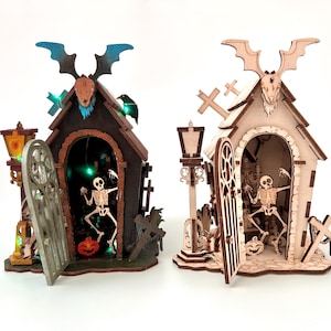 Könnte beinhalten: Zwei Halloween-Holzstrukturen, eine dunkelbraune, eine hellbraune, jeweils mit einer Skelettfigur im Inneren. Beide haben Fledermausdekorationen, offene Türen, einen Kürbis und einen Grabstein. Sie sind mit grünen Lichtern beleuchtet.