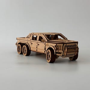 Op de afbeelding: Een houten model van een pick-up met zes wielen. Het model is gemaakt van lichtbruin hout en heeft gedetailleerde uitsparingen voor de ramen, wielen en carrosserie. De truck heeft een grille en een laadbak.