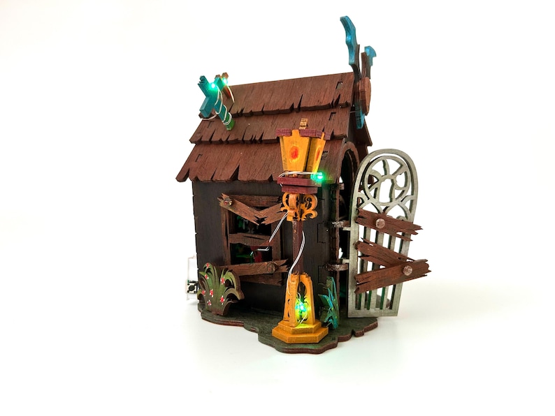 May include: A miniature haunted house with a brown shingled roof, a broken window, and a decorative gate. The house has a lamppost with a glowing green light and is decorated with green and orange lights.