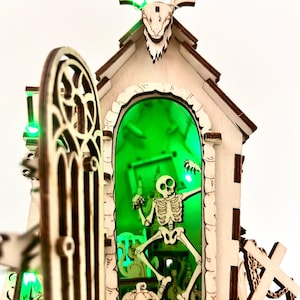 May include: A wooden Halloween-themed decoration featuring a skeleton inside a building with a green-lit interior. The building has a decorative door, a carved bat-like figure on top, and a carved pumpkin. The overall design is intricate and detailed.