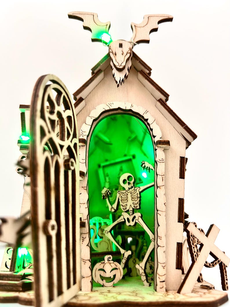 May include: A wooden Halloween-themed decoration featuring a skeleton inside a building with a green-lit interior. The building has a decorative door, a carved bat-like figure on top, and a carved pumpkin. The overall design is intricate and detailed.