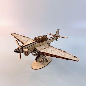Könnte beinhalten: Ein Holzmodellflugzeug mit Propeller und detailliertem Flügeldesign. Das Modell besteht aus hellfarbenem Holz und wird auf einem Holzständer präsentiert. Das Flugzeugmodell ist ein Bastelartikel.
