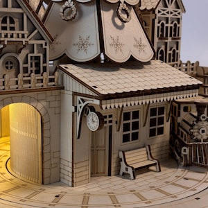 May include: A detailed wooden model of a train station with a clock, bench, and train. The model features intricate cut-out designs, including snowflakes and a decorative wreath. The station has a light inside the archway.