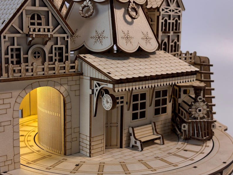 May include: A detailed wooden model of a train station with a clock, bench, and train. The model features intricate cut-out designs, including snowflakes and a decorative wreath. The station has a light inside the archway.