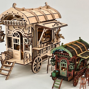 Op de afbeelding: Twee miniatuur houten wagenmodellen met ingewikkelde lasergesneden details. De grotere wagen is van licht natuurlijk hout, terwijl de kleinere is geschilderd in groen-, rood- en bruintinten. Beide bevatten kleine lantaarns, trappen en krukjes.