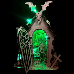 May include: Wooden Halloween decoration featuring a spooky church with a skeleton inside, illuminated by green lights. The church has a bat-shaped roof and a decorative door. A pumpkin and a cross are also visible.