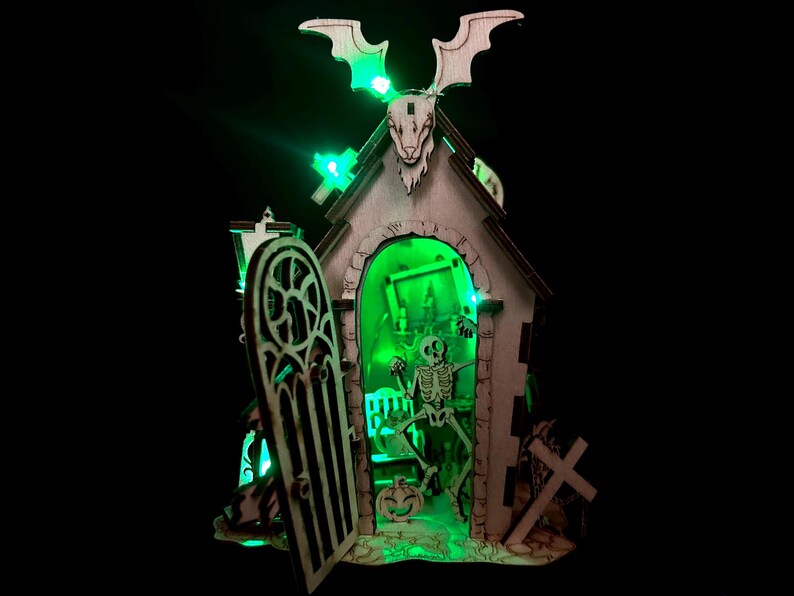 May include: Wooden Halloween decoration featuring a spooky church with a skeleton inside, illuminated by green lights. The church has a bat-shaped roof and a decorative door. A pumpkin and a cross are also visible.