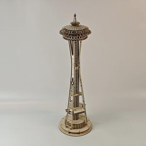 May include: A wooden model of the Seattle Space Needle. The model is made of light-colored wood and features intricate details, including the observation deck and spire. The base is circular, and the tower is tall and slender.
