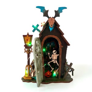 May include: A Halloween-themed miniature house with a skeleton inside, a pumpkin, and a decorative lamp post. The house has a bat-like figure on top and a cross. The scene is illuminated with green lights.