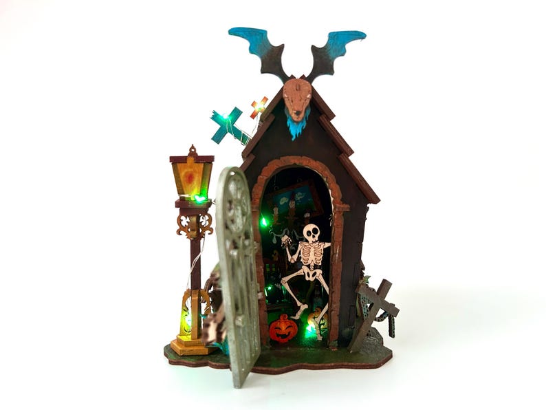 May include: A Halloween-themed miniature house with a skeleton inside, a pumpkin, and a decorative lamp post. The house has a bat-like figure on top and a cross. The scene is illuminated with green lights.