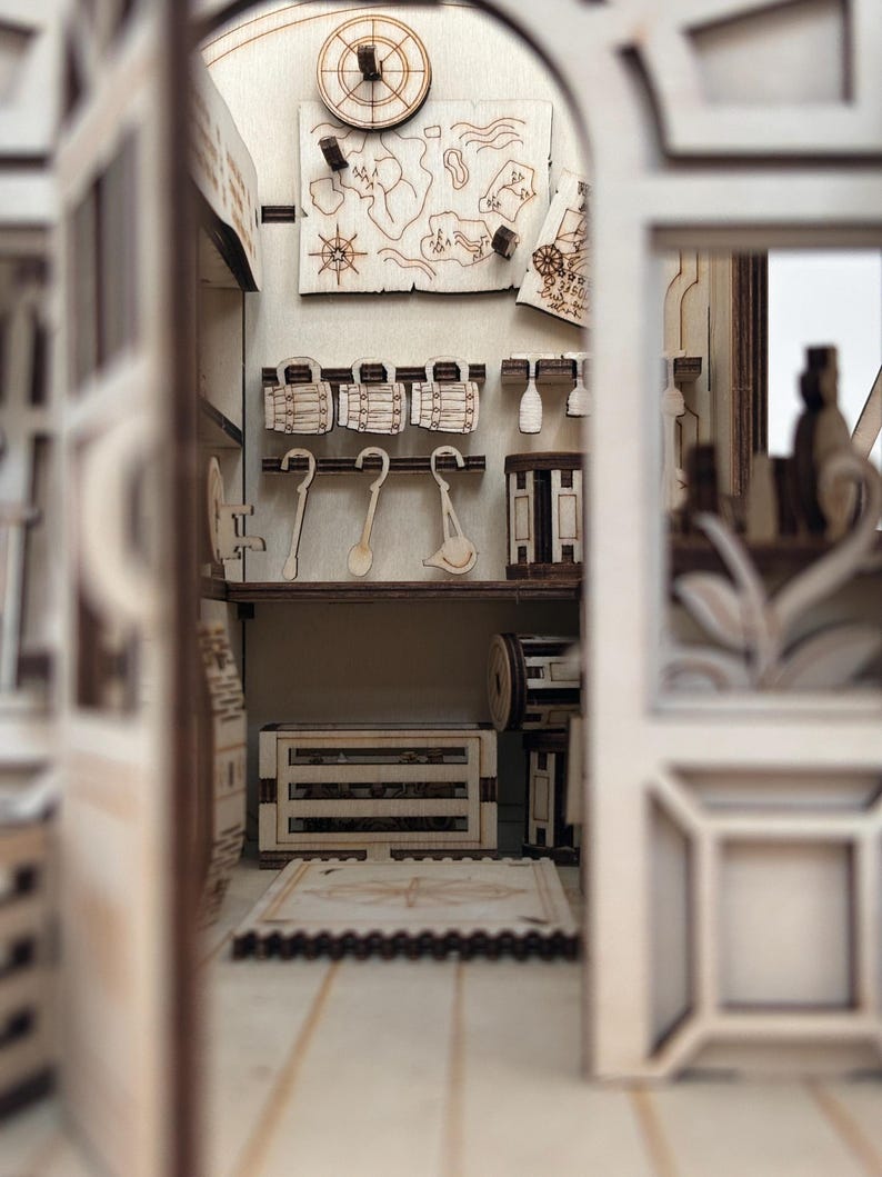 Puede incluir: Un diorama de madera en miniatura de un interior de taberna con detalles cortados con l&aacute;ser. Las paredes muestran un mapa, jarras colgantes y cucharones. Una caja de madera se asienta en el suelo cerca de una alfombra con dise&ntilde;o de br&uacute;jula.
