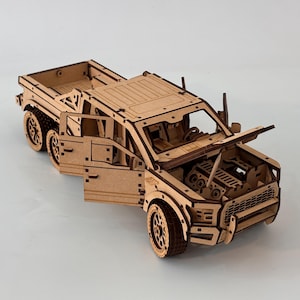 May include: A detailed wooden model of a pickup truck with six wheels. The doors and hood are open, revealing the interior and engine. The model is made of light brown wood and sits on a white surface.