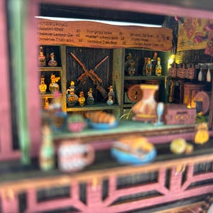 Puede incluir: Un diorama de taberna en miniatura que presenta una barra de madera con estantes llenos de botellas peque&ntilde;as, dos rifles cruzados en la pared trasera y un men&uacute; titulado HOUSE DRINKS. La estructura tiene un marco exterior rosa y comida.