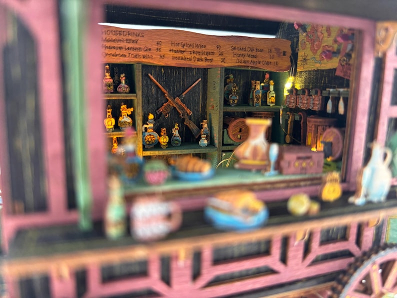 Puede incluir: Un diorama de taberna en miniatura que presenta una barra de madera con estantes llenos de botellas peque&ntilde;as, dos rifles cruzados en la pared trasera y un men&uacute; titulado HOUSE DRINKS. La estructura tiene un marco exterior rosa y comida.