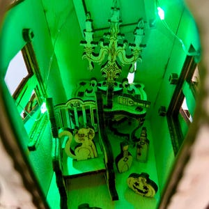 May include: A miniature wooden haunted house with a green interior illuminated by LED lights. The interior features a chandelier, furniture, and decorative wooden cutouts of a squirrel, pumpkins, and bottles. The overall theme is Halloween.