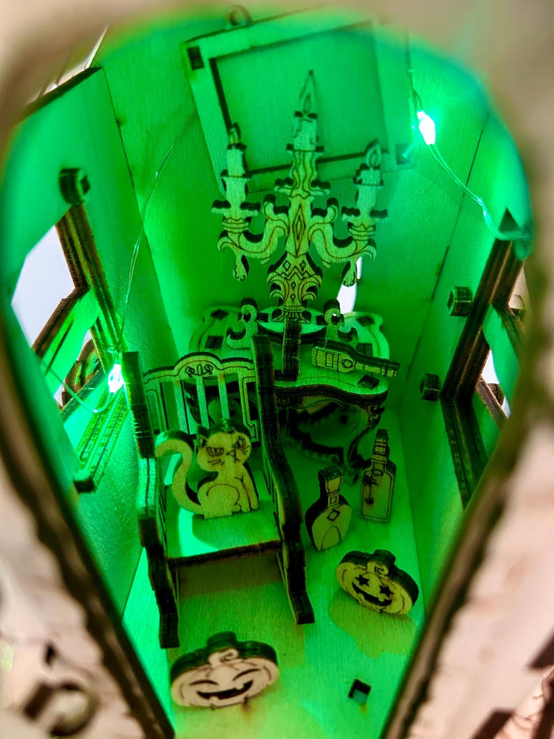 May include: A miniature wooden haunted house with a green interior illuminated by LED lights. The interior features a chandelier, furniture, and decorative wooden cutouts of a squirrel, pumpkins, and bottles. The overall theme is Halloween.