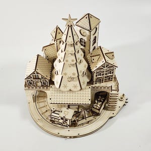 May include: A wooden Christmas village model with a central Christmas tree, houses, a clock tower, and a train track. The model is made of light-colored wood and features intricate cut-out details.