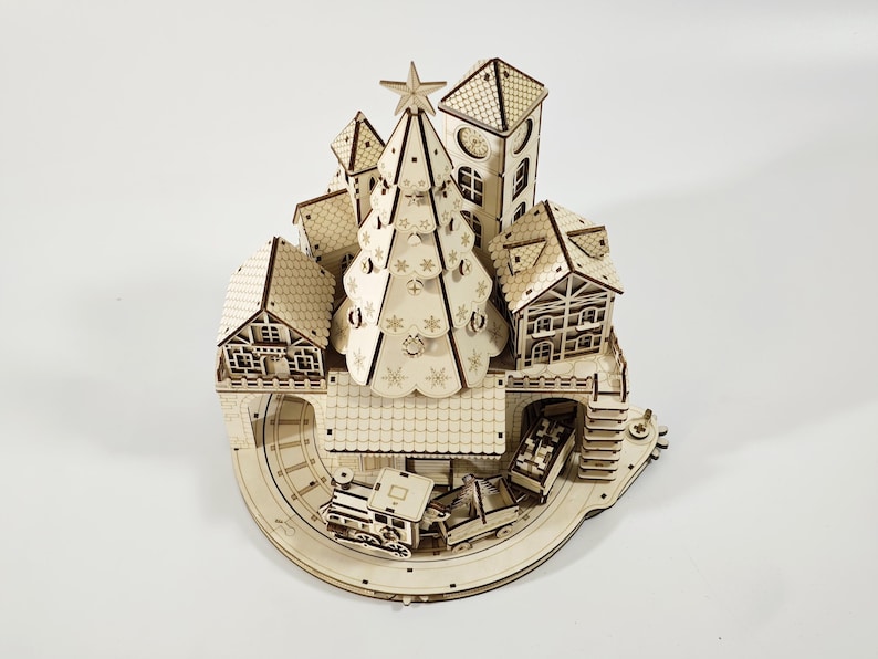 May include: A wooden Christmas village model with a central Christmas tree, houses, a clock tower, and a train track. The model is made of light-colored wood and features intricate cut-out details.