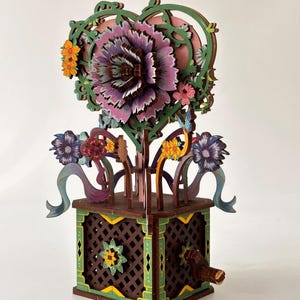 Puede incluir: Una caja de música de madera con una gran flor morada y rosa en capas en la parte superior, rodeada de enredaderas verdes y un marco en forma de corazón. La caja tiene un diseño de celosía marrón con detalles florales verdes y amarillos.