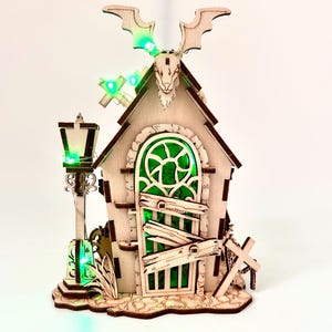 May include: A wooden Halloween-themed house with a green-tinted window and door, a bat-shaped decoration, and a lamppost. The house has a cross and a boarded-up door. Green LED lights illuminate the scene.