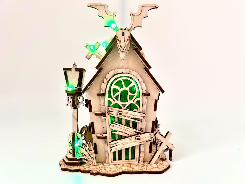 May include: A wooden Halloween-themed house with a green-tinted window and door, a bat-shaped decoration, and a lamppost. The house has a cross and a boarded-up door. Green LED lights illuminate the scene.