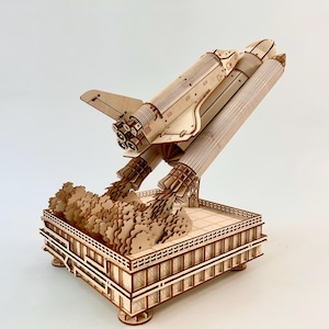 May include: A light-colored wooden model of a space shuttle launching from a rectangular platform. The shuttle is angled upward with detailed thrusters and wings, while layered wooden cutouts represent exhaust clouds rising from the base.