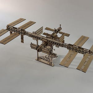 Op de afbeelding: Een lasergesneden houten model van het Internationaal Ruimtestation met een centrale vakwerkstructuur, meerdere rechthoekige zonnepaneelvleugels en diverse modulaire onderdelen, ondersteund door een stevige standaard op een neutrale ondergrond.
