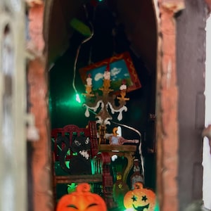 May include: A miniature Halloween-themed diorama featuring a brick archway. Inside, a black cat sits on a red chair, with a small painting and a candelabra. Two carved orange pumpkins with smiling faces are in the foreground.