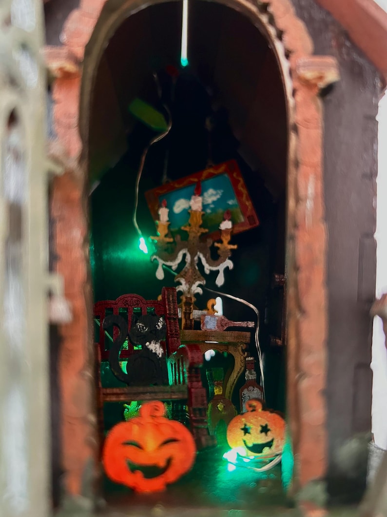 May include: A miniature Halloween-themed diorama featuring a brick archway. Inside, a black cat sits on a red chair, with a small painting and a candelabra. Two carved orange pumpkins with smiling faces are in the foreground.