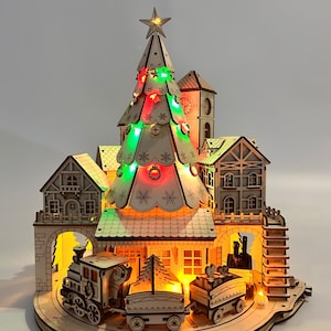 May include: A wooden Christmas village scene with a lit-up Christmas tree, buildings, and a train. The tree is adorned with red and green lights and topped with a star. The train has three cars and is traveling around the village. The scene is illuminated with warm lights.