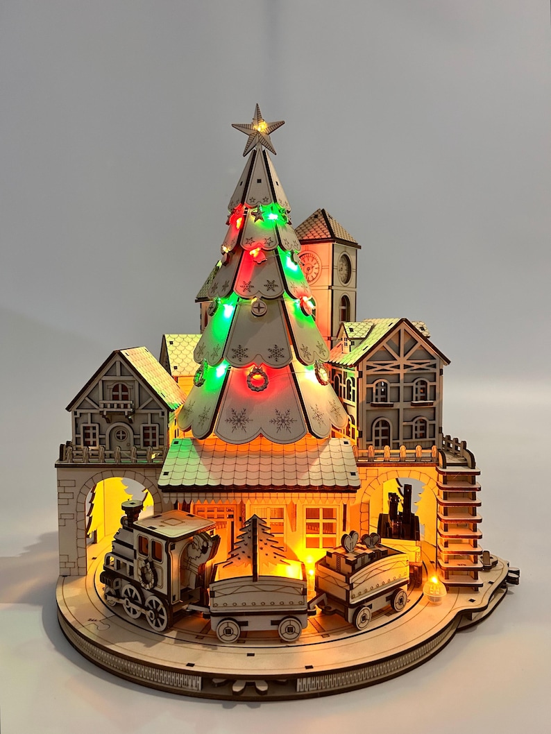 May include: A wooden Christmas village scene with a lit-up Christmas tree, buildings, and a train. The tree is adorned with red and green lights and topped with a star. The train has three cars and is traveling around the village. The scene is illuminated with warm lights.